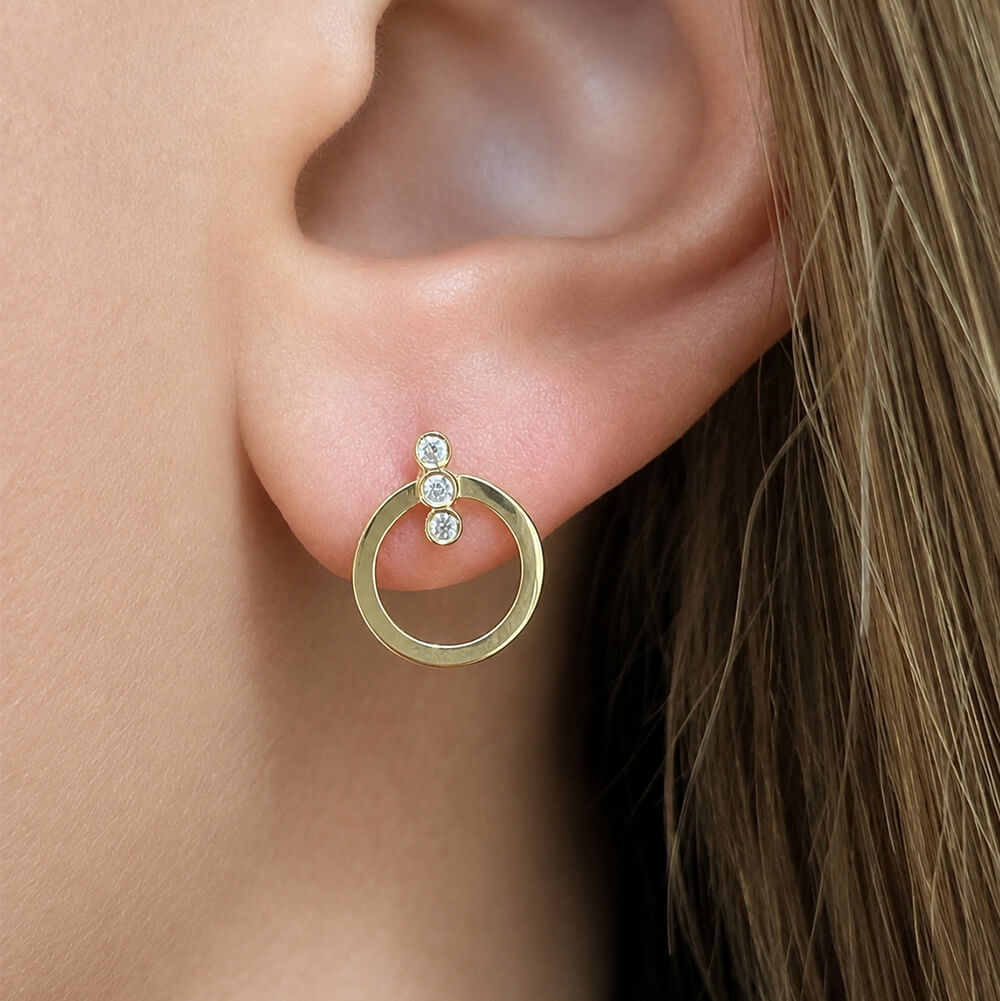 A close-up of a woman's ear wearing a gold hoop earring with three small clear diamonds stacked vertically on the post.