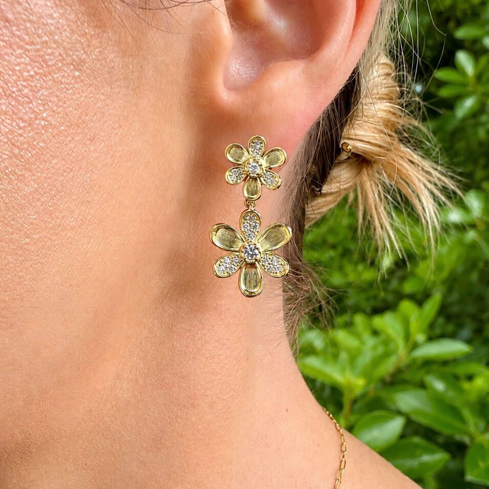 A close-up of a woman's ear wearing a pair of gold flower-shaped drop earrings with clear diamonds, set against a blurred green outdoor background.