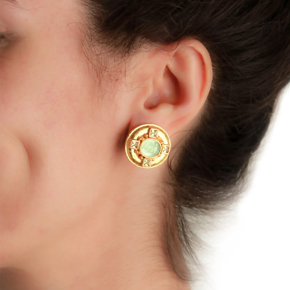 A close-up of a woman's ear wearing a round gold stud earring with a light green oval center stone.