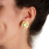 A close-up of a woman's ear wearing a round gold stud earring with a light green oval center stone.