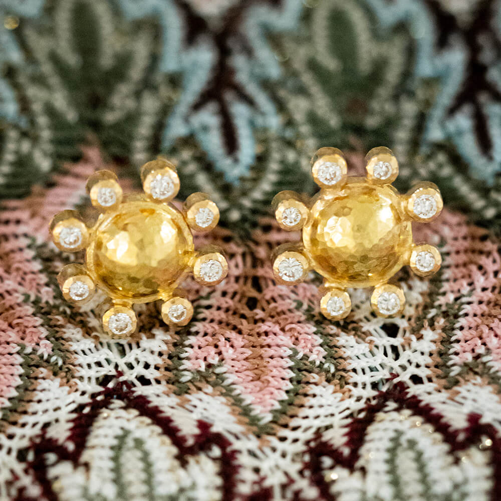 A pair of Elizabeth Locke 19K yellow gold sunburst stud earrings with small clear diamonds, displayed on a textured pink, green, and white lace fabric.