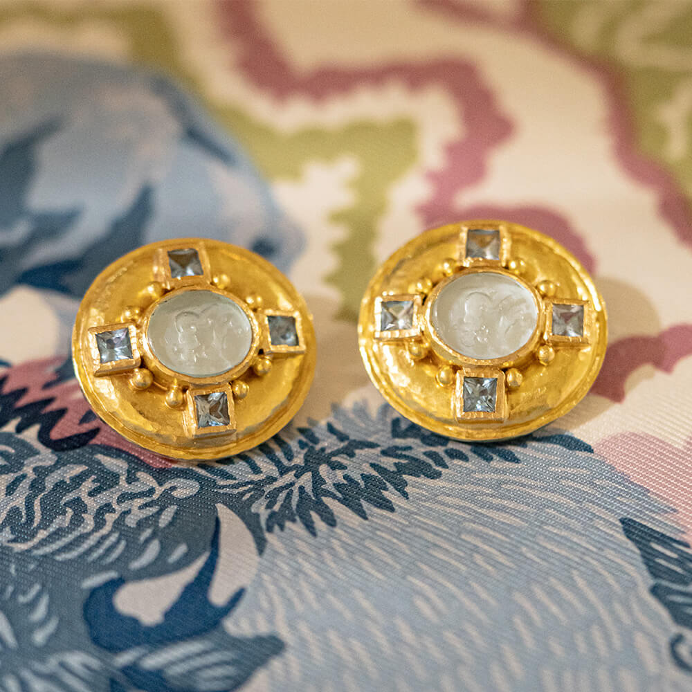 A pair of round 19k yellow gold earrings with central ligth aqua venetian intaglio stones and four small square blue stones around the edge, displayed on a patterned fabric surface.