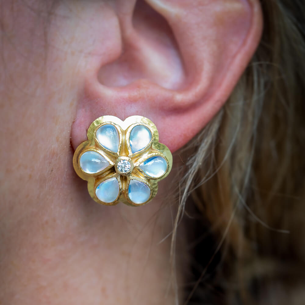 A close-up of a 19K yellow gold flower-shaped stud earring with pale blue moonstone inlay petals and a small central clear diamond, worn on a person's ear.