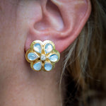 A close-up of a 19K yellow gold flower-shaped stud earring with pale blue moonstone inlay petals and a small central clear diamond, worn on a person's ear.
