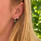 A close-up of a woman's ear wearing a 14K yellow gold teardrop stud earring with a green emerald center stone, alongside a smaller clear gemstone stud, with her blonde hair and a blurred green outdoor background.