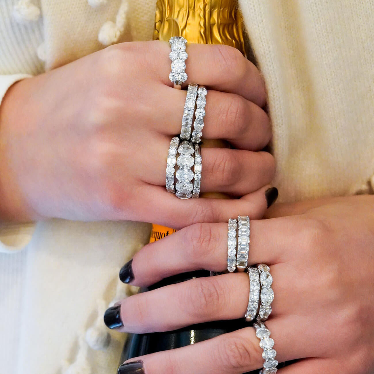 Close-up of a woman's hands wearing multiple platinum eternity rings set with diamonds, styled with a cream knit sweater and a gold bottle in the background.