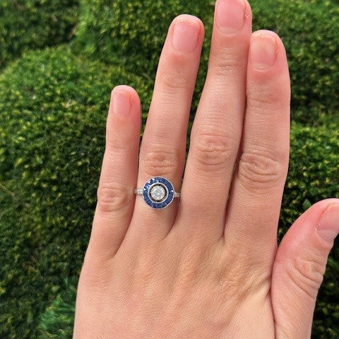 Hand wearing a Bulls Eye Diamond and Sapphire ring against a green moss background