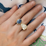 A close-up of a hand wearing an Estate 18K yellow gold rings with a deep blue sapphire and a pear-shaped diamond, set against a patterned fabric background.