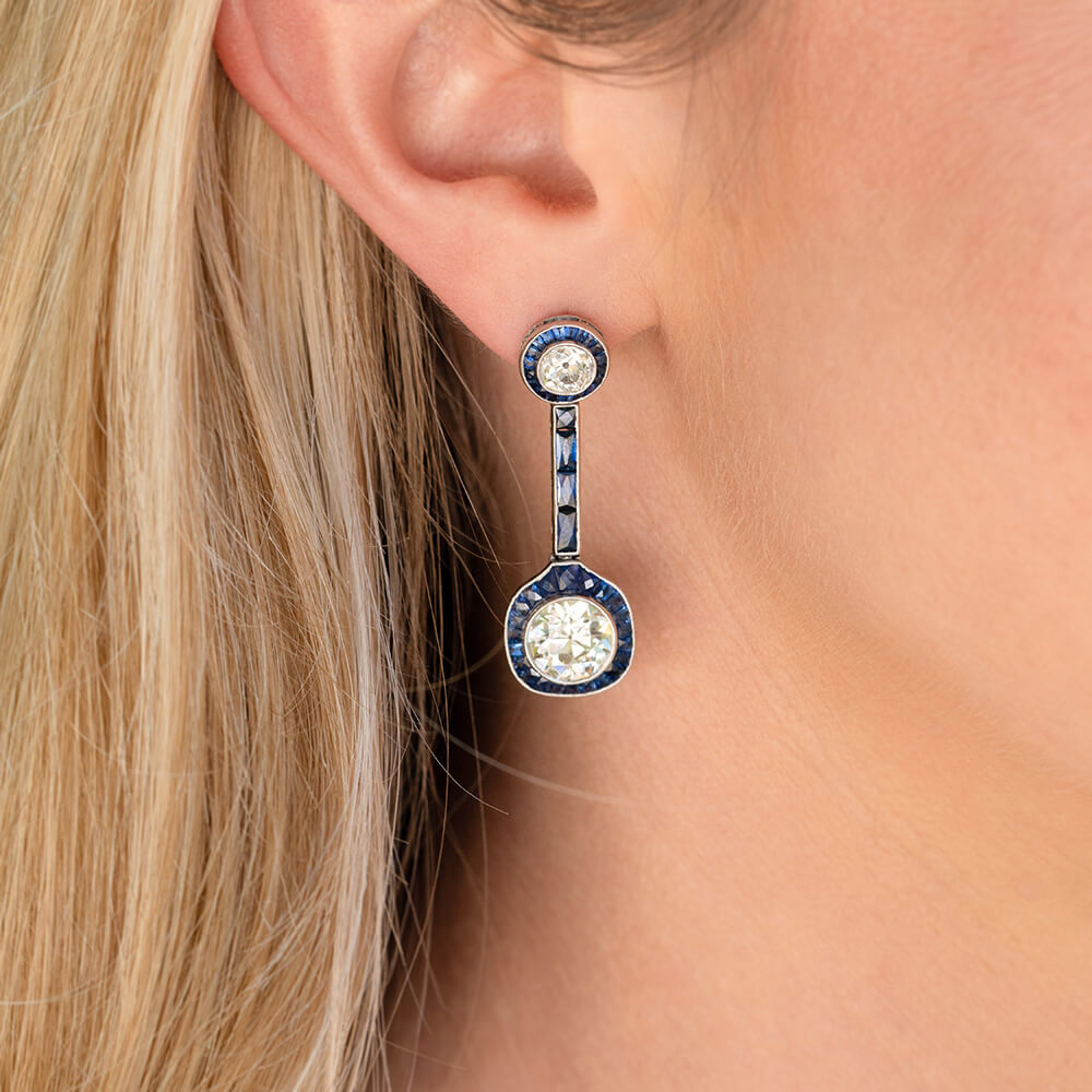 A close-up of a model's ear wearing a single drop earring. The earring features a round diamond with sapphire halo at the  top and a larger matching round diamond with sapphire halo at the bottom, 