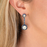 A close-up of a model's ear wearing a single drop earring. The earring features a round diamond with sapphire halo at the  top and a larger matching round diamond with sapphire halo at the bottom, 