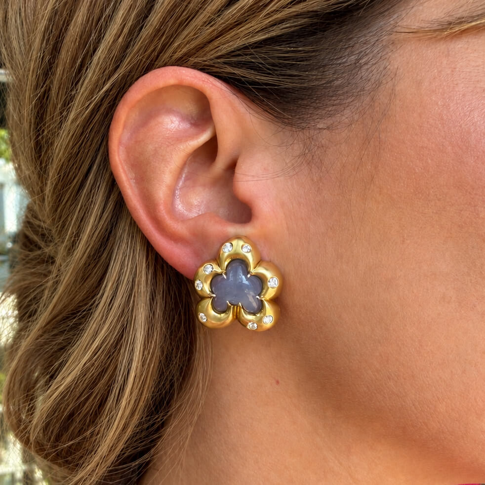 A close-up of a woman's ear wearing a gold flower-shaped stud earring with a gray-blue moonstone in the center and small clear diamond accent stones.