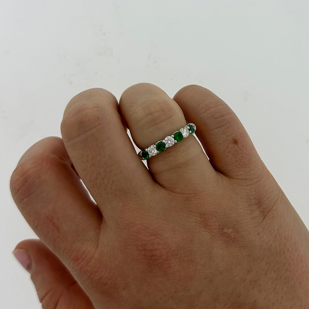A close-up of a hand wearing a platinum band ring set with alternating green emeralds and clear round diamonds, against a plain light background.