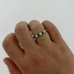 A close-up of a hand wearing a platinum band ring set with alternating green emeralds and clear round diamonds, against a plain light background.