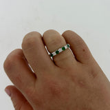 A close-up of a hand wearing a platinum band ring set with alternating green emeralds and clear round diamonds, against a plain light background.