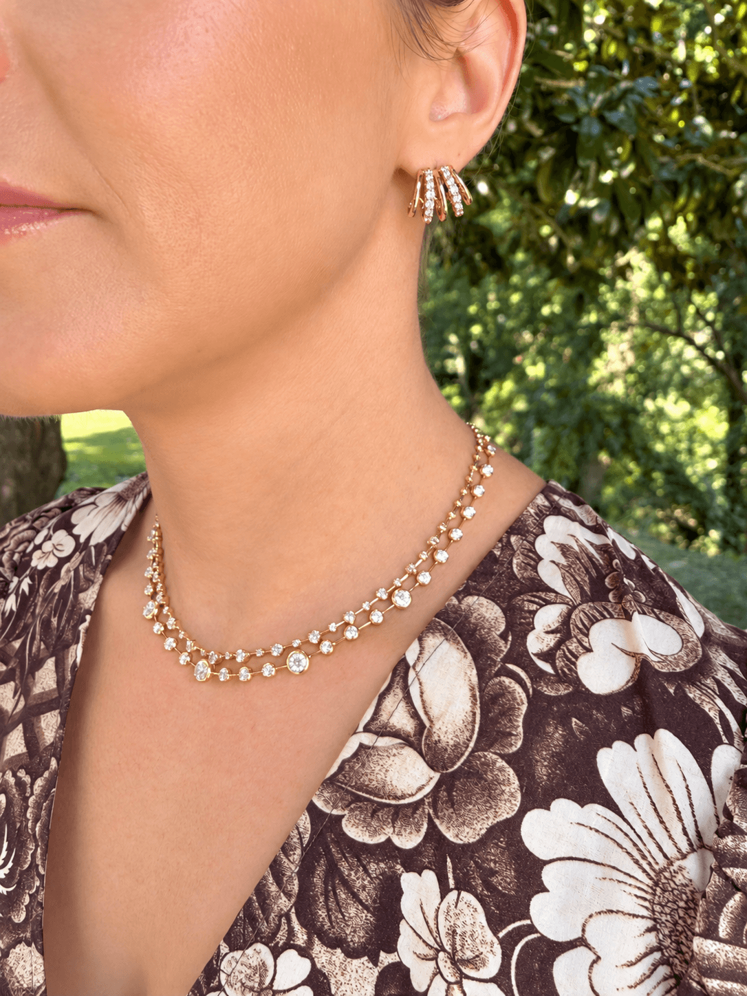 A close-up lifestyle shot of a woman wearing a delicate gold necklace with small clear stones and matching gold hoop earrings, set against a blurred green outdoor background.