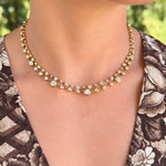A close-up of a model wearing a delicate 18K yellow gold double-strand necklace adorned with small clear round-cut diamonds, set against a blurred green outdoor background.