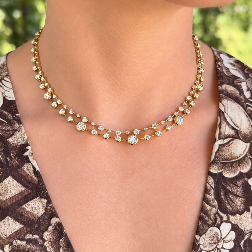 A close-up of a model wearing a delicate 18K yellow gold double-strand necklace adorned with small clear round-cut diamonds, set against a blurred green outdoor background.
