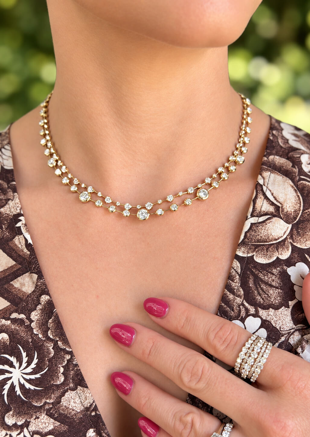 A close-up of a model wearing a Kwiat 18K yellow gold and diamond necklace, with Kwiat diamond rings on her fingers and a brown floral-patterned top in the background.