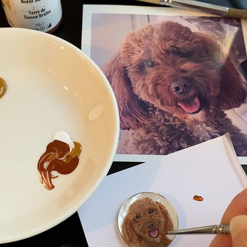 A close-up of a hand painting a small round pet portrait of a brown poodle dog, with a reference photo of the dog, a white paint palette with brown and white paint, and Golden Fluid Acrylic paint cans in the background.