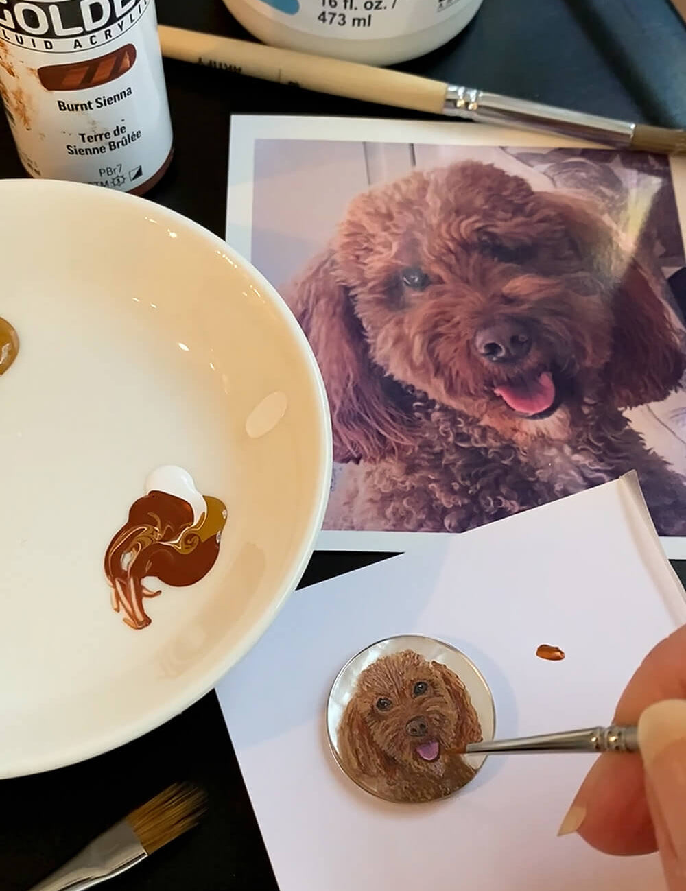 A close-up of a hand painting a small round pet portrait of a brown poodle dog, with a reference photo of the dog, a white paint palette with brown and white paint, and Golden Fluid Acrylic paint cans in the background.