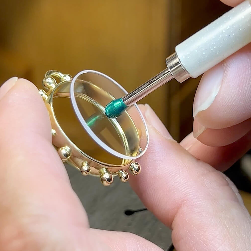A close-up of a person's hands holding a gold ring under a magnifying lens while applying a small amount of green adhesive or polish with a white-handled tool.