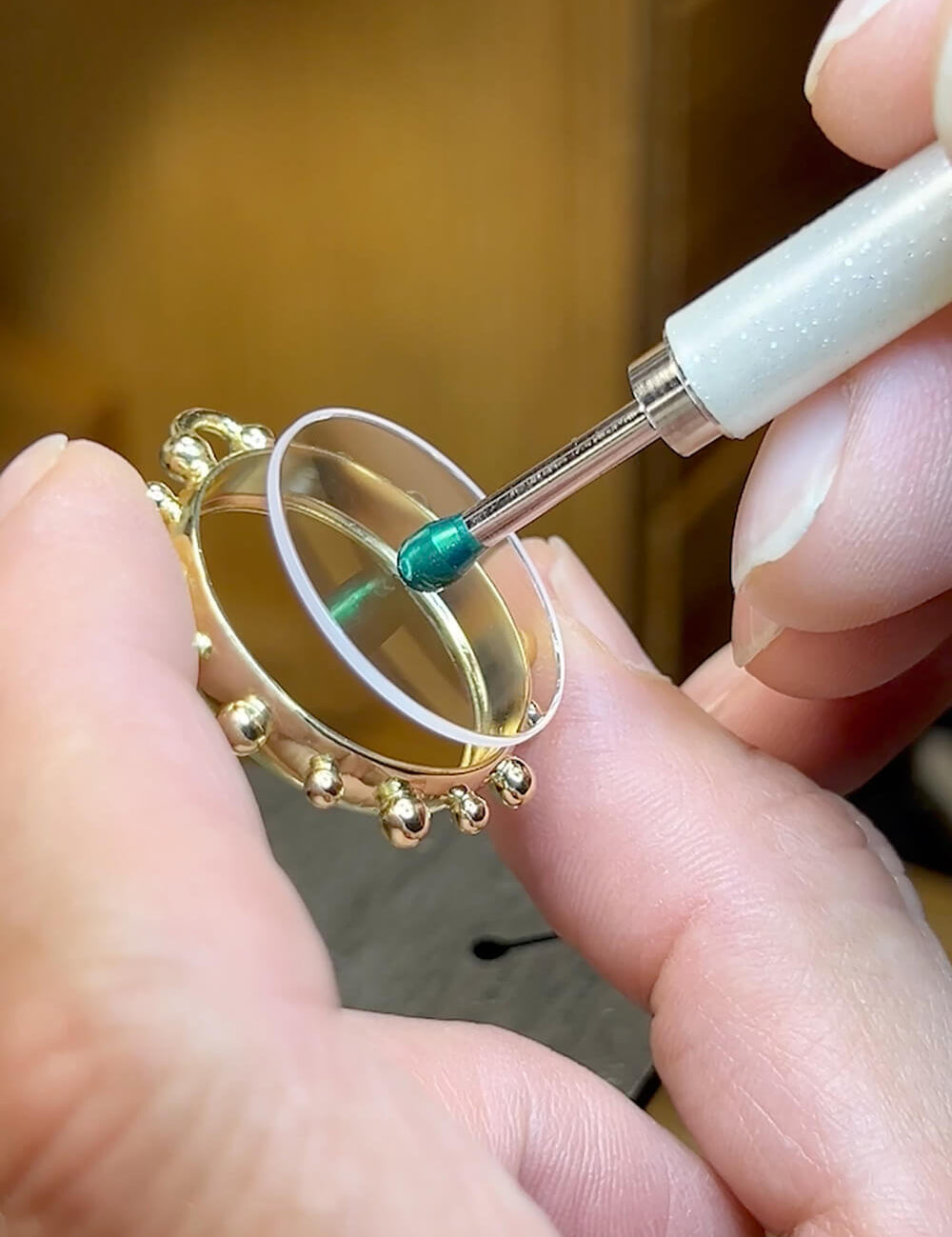 A close-up of a person's hands holding a gold ring under a magnifying lens while applying a small amount of green adhesive or polish with a white-handled tool.