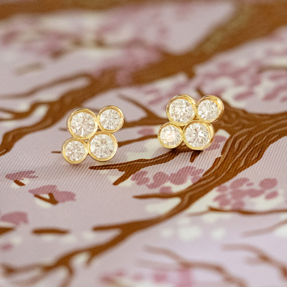 A pair of 18K yellow gold stud earrings with four round clear diamonds each, displayed on a pink and brown cherry blossom patterned fabric.