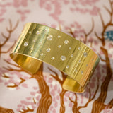 A wide 18K yellow gold cuff bracelet with a brushed finish and small clear diamonds photographed on a pink and brown cherry blossom patterned surface.