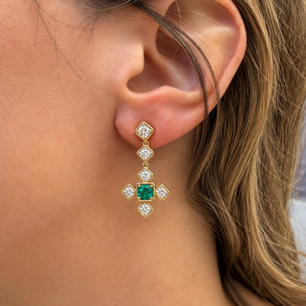A close-up of an 18k yellow gold dangle earring worn on a woman's ear, featuring a central emerald stone surrounded by clear diamond stones in a cross-like design.