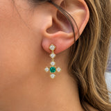 A close-up of an 18k yellow gold dangle earring worn on a woman's ear, featuring a central emerald stone surrounded by clear diamond stones in a cross-like design.