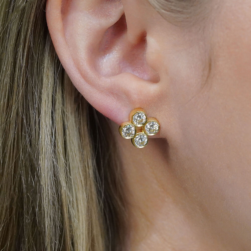 A close-up of a woman's ear wearing an 18K yellow gold stud earring with four clear diamonds in a quad design cluster.