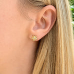 A close-up of a woman's ear wearing a small 18K yellow gold flower-shaped stud earring with a center diamond