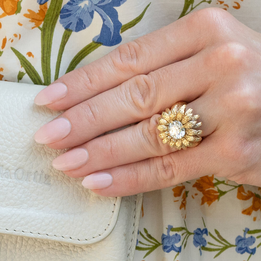 A close-up of a hand wearing a gold ring with a large clear center stone in a sunburst flower design, resting on a white textured leather clutch with a floral print background.