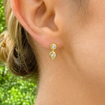 A close-up of a woman's ear wearing a 14K gold earring with two diamonds, set against a blurred green outdoor background.