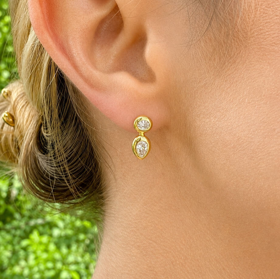 A close-up of a woman's ear wearing a 14K gold earring with two diamonds, set against a blurred green outdoor background.