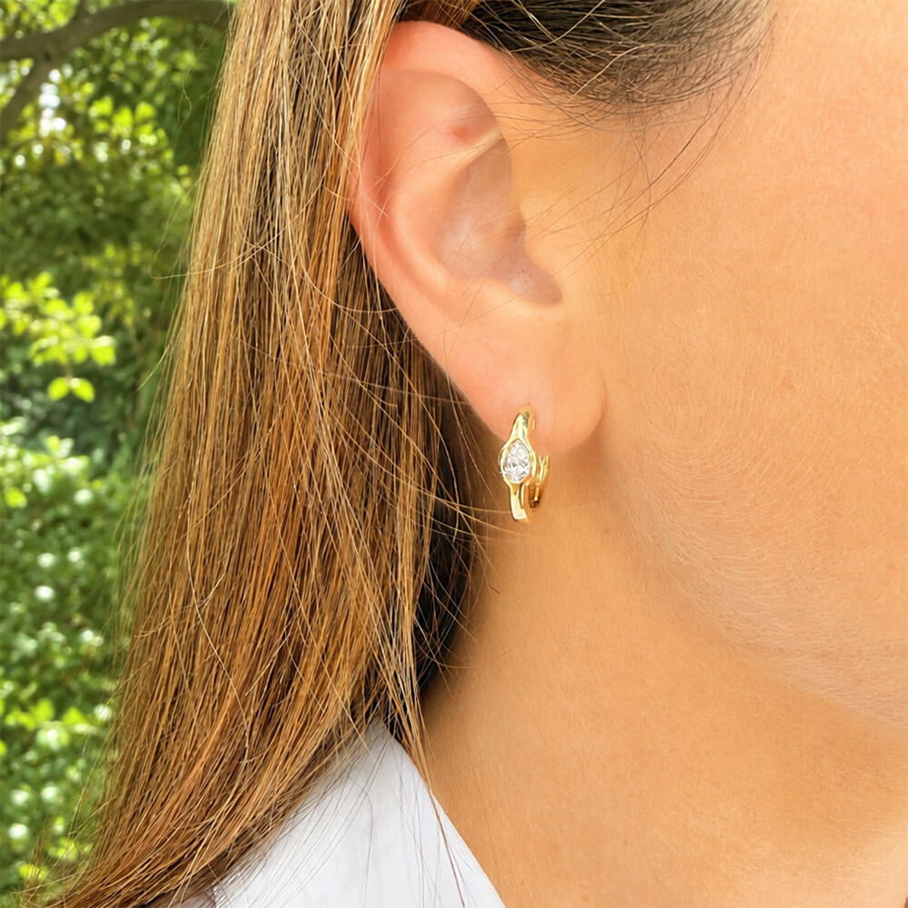 A close-up of a woman's ear wearing a small 14K gold huggie hoop earring with a clear pear-cut diamond, set against a blurred green outdoor background.