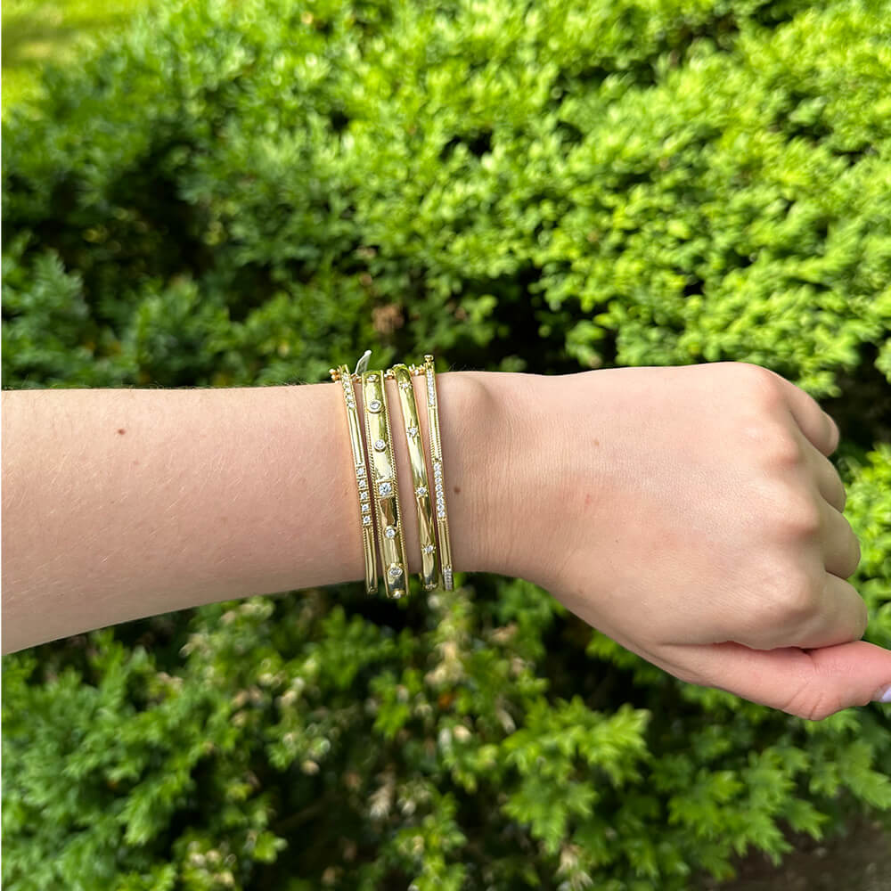Hand wearing multiple gold bracelets with a green bush background