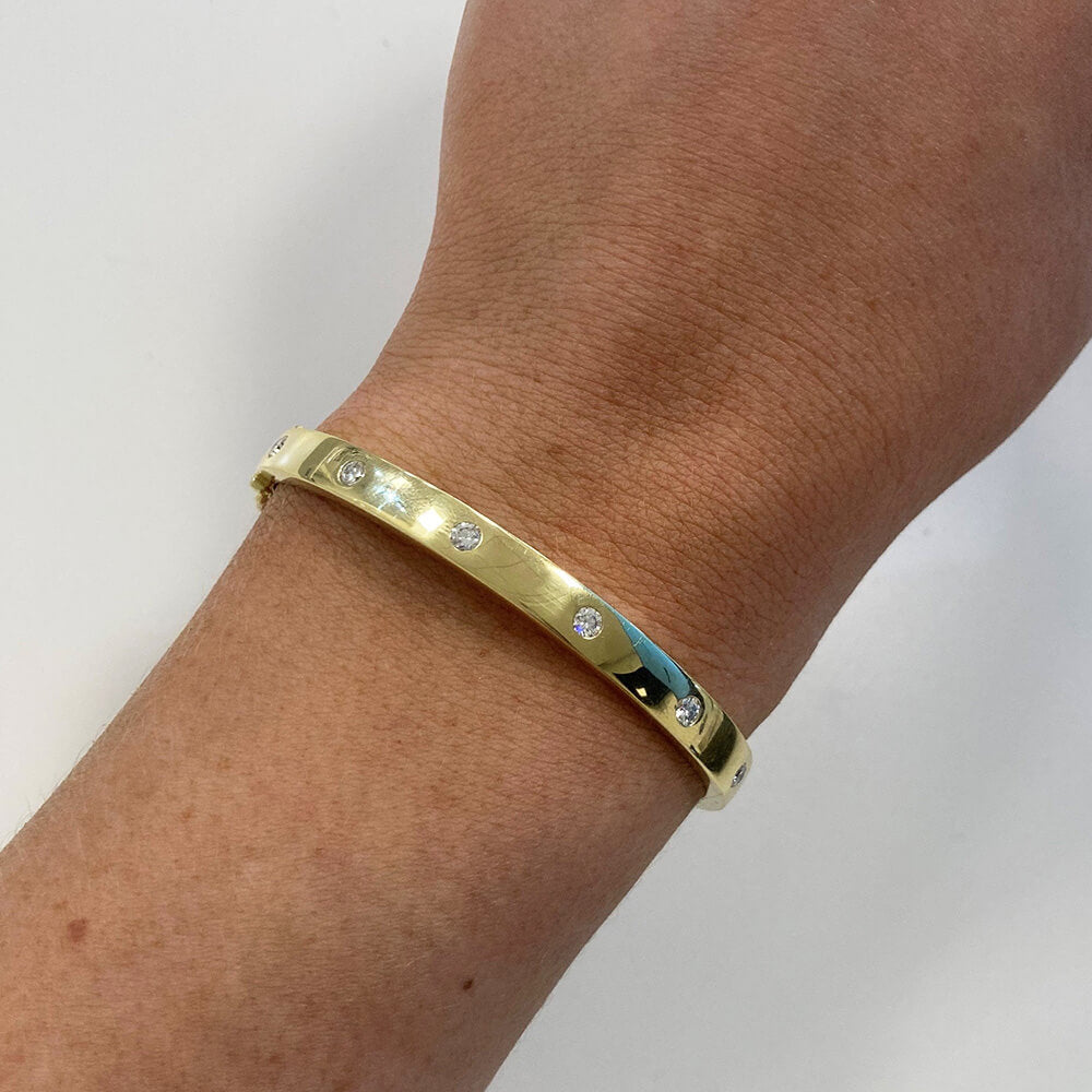 An 18K yellow gold bangle bracelet with small inset clear diamonds, shown worn on a woman's wrist against a plain light background.