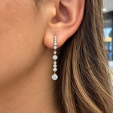 A close-up of a woman's ear wearing a long drop earring made of clear diamonds in an 18k white gold setting. The earring features multiple graduated round stones hanging vertically.
