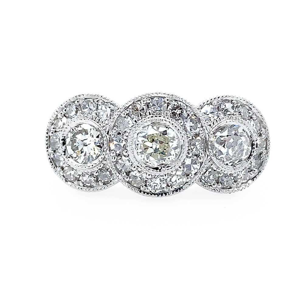 A platinum three-stone bullseye diamond ring set against a plain white background. Each round diamond  is surrounded by a halo of smaller round diamonds, with a milgrain-style border.