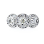 A platinum three-stone bullseye diamond ring set against a plain white background. Each round diamond  is surrounded by a halo of smaller round diamonds, with a milgrain-style border.