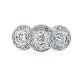 A platinum three-stone bullseye diamond ring set against a plain white background. Each round diamond  is surrounded by a halo of smaller round diamonds, with a milgrain-style border.