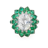 A vintage-style floral inspired ring featuring a large oval center diamond surrounded by a radiant halo of emeralds and small accent diamonds in platinum metal, set against a plain white background.