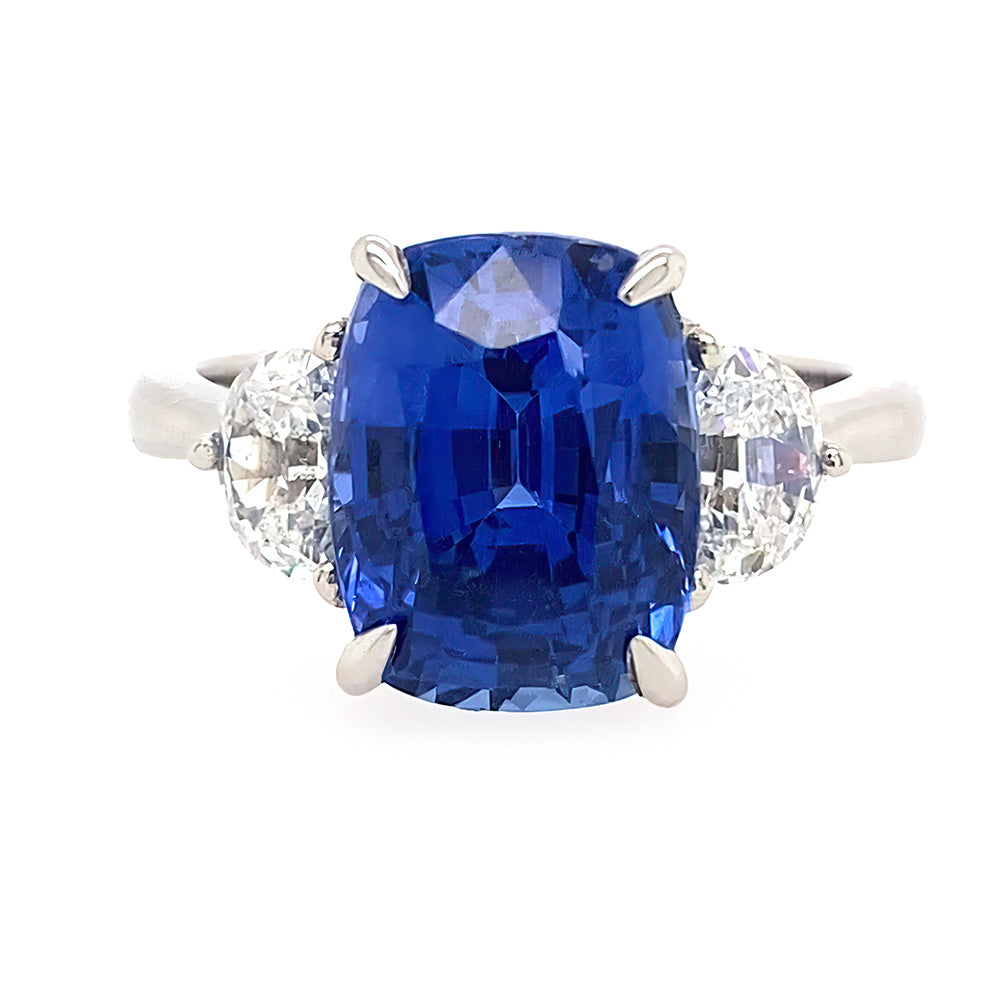 A three-stone ring featuring a large cushion-cut blue sapphire center stone flanked by two half moon clear diamonds in a platinum setting, shown against a plain white background.