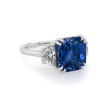 A platinum ring featuring a large, square-cut deep blue sapphire center stone, accented by two small half moon-cut diamonds on either side of the band, set against a clean white background.