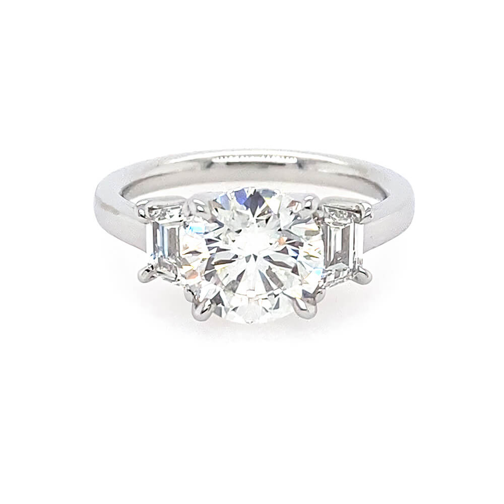 A three-stone diamond engagement ring with a round center diamond and trapezoid side diamonds on a polished platinum band, set against a plain white background.