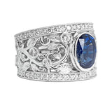 An ornate platinum ring featuring a large oval-cut blue sapphire center stone, surrounded by intricate filigree leaf and scroll motifs, round diamonds, and a diamond-accented border, shown on a plain white background.