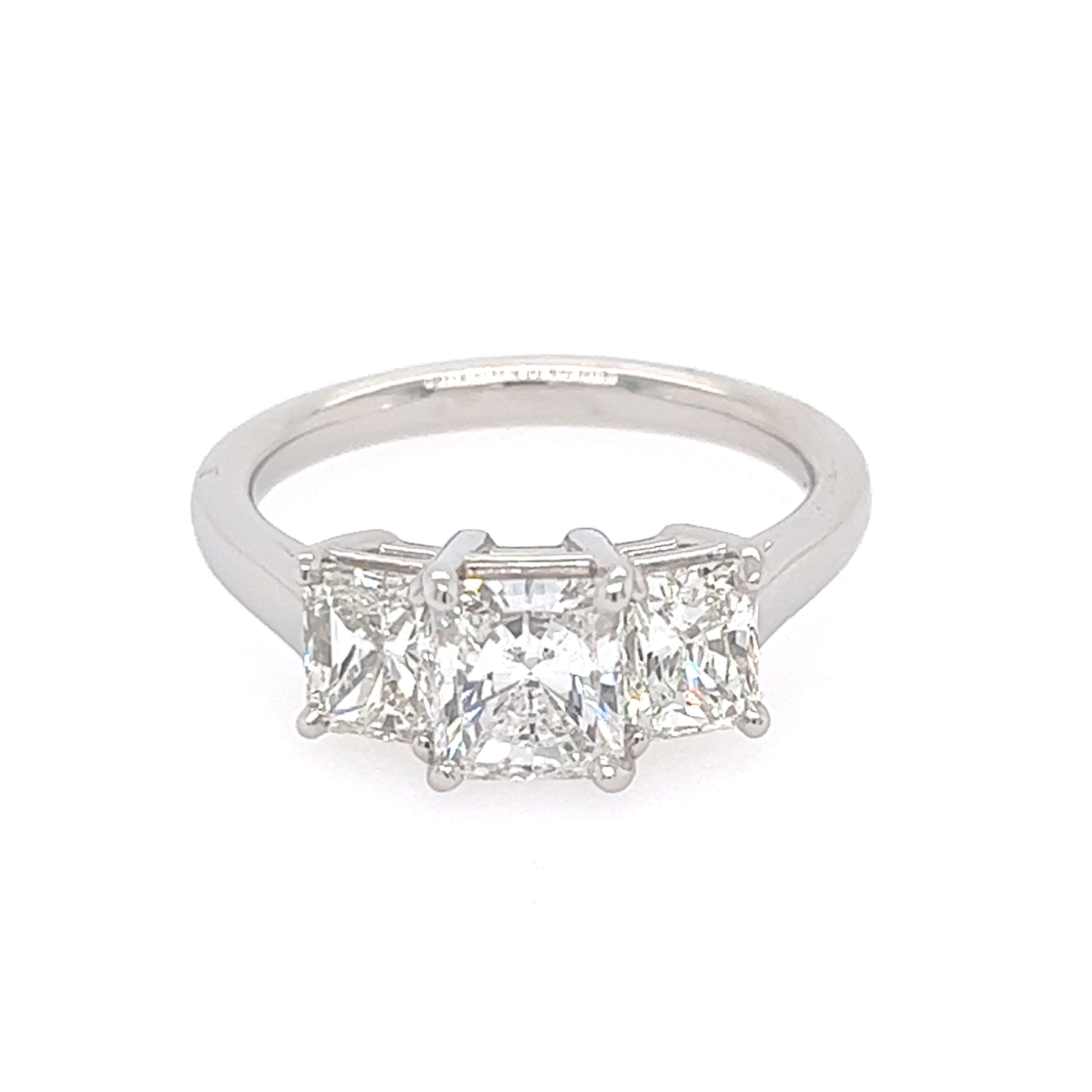 This image features an elegant Platinum and Diamond Three Stone Ring, presented against a simple white background that highlights its intricate details. The ring showcases a classic design with three prominently set radiant-cut diamonds, each encased in a sleek, polished setting.