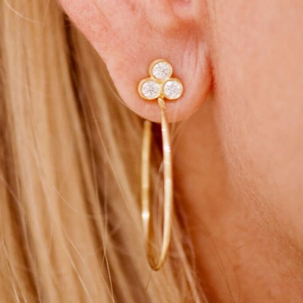 Close-up of an 18K gold earring with a clover design worn by a person with blonde hair.