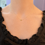 A delicate gold chain necklace with a small gold heart pendant, shown worn on a model's neck over a black lace-trimmed top.
